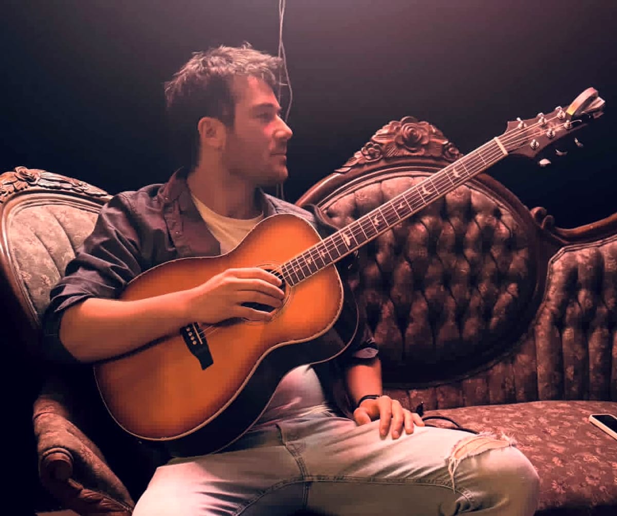 Nate Couture, guitare en main, assis dans un fauteuil ancien